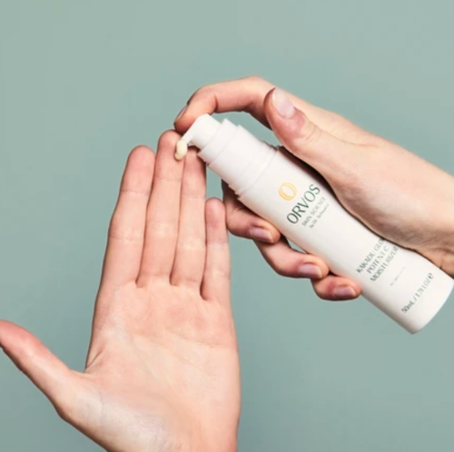 A close-up of hands dispensing a white cream from an ORVOS cosmetic pump bottle against a muted sage green background.