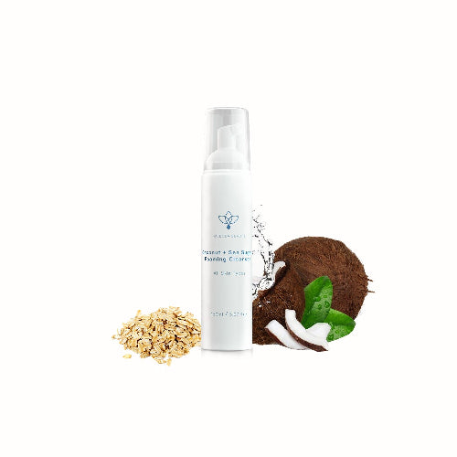 A white pump bottle of Coconut + Sea Sulfur Cleansing Cream centered on a white background, with small ingredient props including oats, a coconut half, and green leaves beside it.