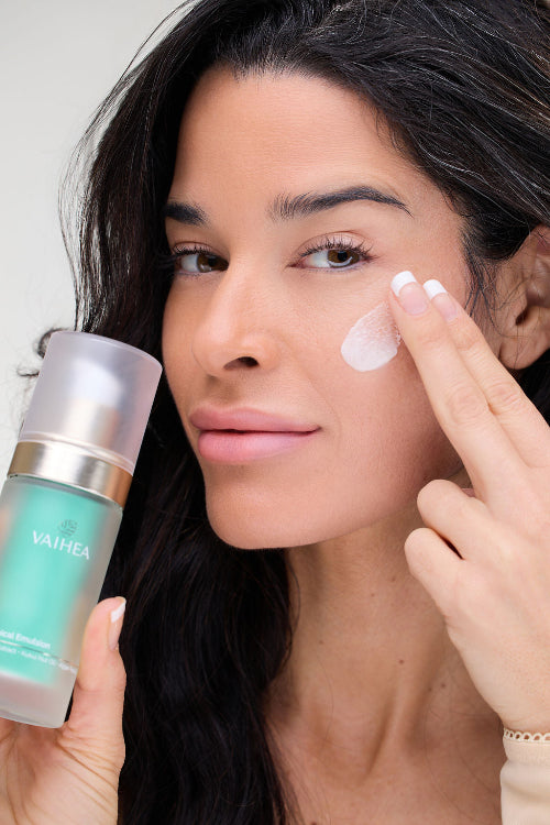Woman applying cream to her face with a bottle of VAIHEA skincare product.