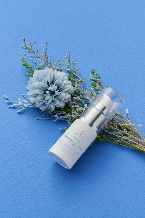 A white skincare pump bottle with a clear cap lies diagonally on a textured blue background, styled with dried lavender and a pale blue flower.