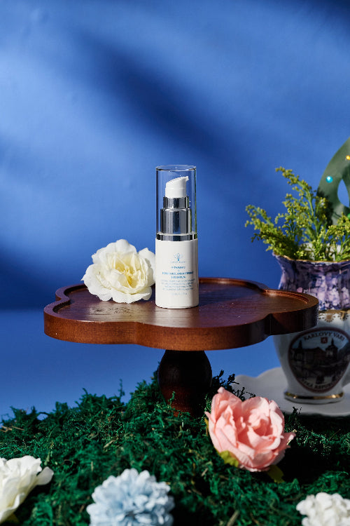 A skincare pump bottle with a clear cap stands on a small wooden pedestal, surrounded by flowers and greenery against a blue backdrop.