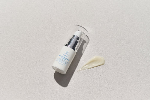 A white cosmetic pump bottle with blue text lies on a light textured surface next to a smear of pale cream product. No person, animal, or real environment is present, so this is Product/Packaging.