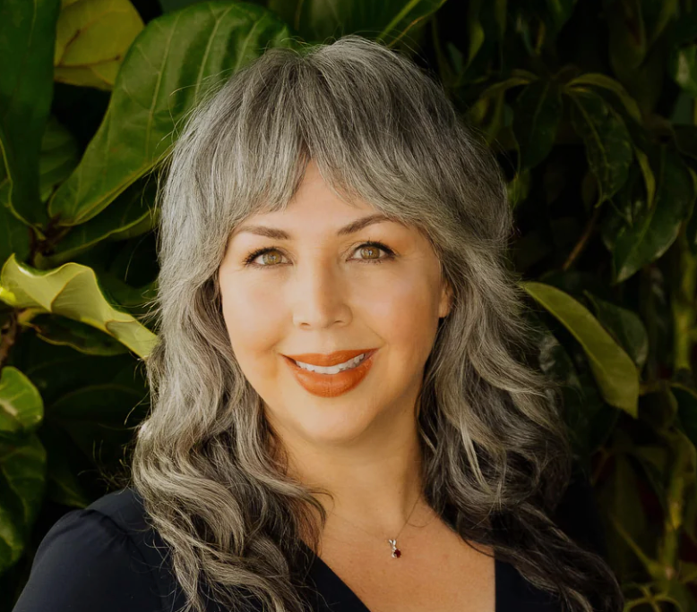 Woman with gray hair smiling in front of green foliage.