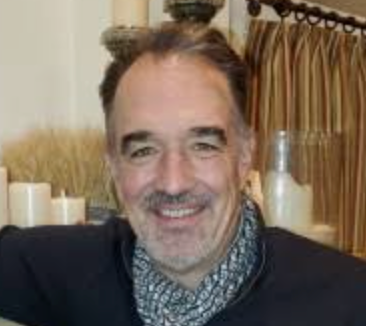 Person with a beard and long hair, wearing a dark sweater and patterned scarf, smiling indoors.