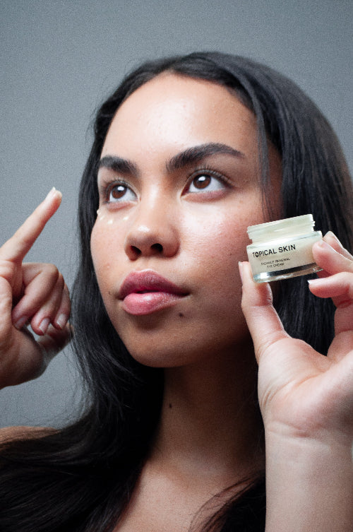 Woman holding a jar of 'Topical Skin' cream near her face against a gray background