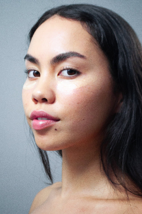 A close-up portrait of a woman with dark hair and natural skin texture, shown from the shoulders up against a plain gray background.