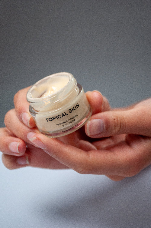 A person's hands holding a small clear jar of TOPICAL SKIN Radiant Renew Eye Cream against a neutral gray background.