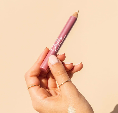 A hand holding a pink shimmer highlighter pencil with a beige tip, set against a solid light beige background. The pencil features white script text that reads 'Chella'