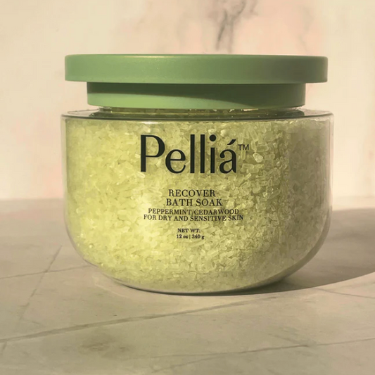 A clear plastic jar of Pellía Recover Bath Soak with a muted sage green lid, filled with pale yellow-green crystalline bath salts, set against a light marble background.