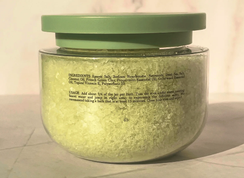 A clear plastic jar filled with pale yellow bath salts, featuring a muted sage green screw-on lid and a white label with black text, set against a neutral light background.