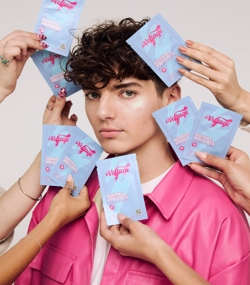 A person in a pink shirt surrounded by multiple hands holding light blue and pink Voltage pimple patch treatment packets against a plain light background.