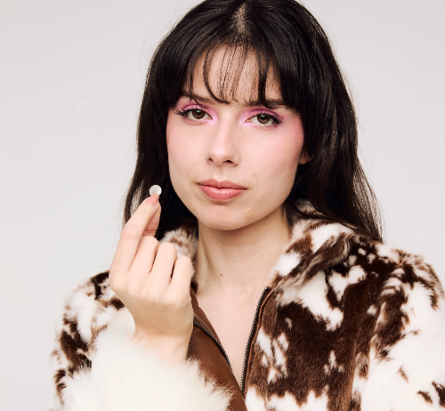 A close-up portrait of a woman with dark hair and pink eye makeup, wearing a brown and white cow-print faux fur zip-up jacket, holding a small white pimple patch between her fingers against a light gray background.