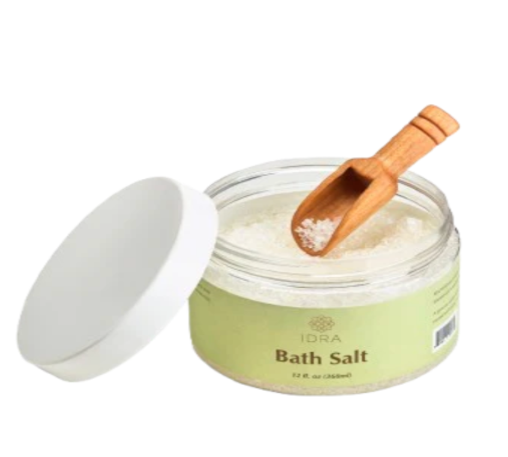 An open jar of IDRA Bath Salt with its white lid leaning beside it, showing white bath salt inside and a small wooden scoop resting in the jar. The label is pale green with black text.