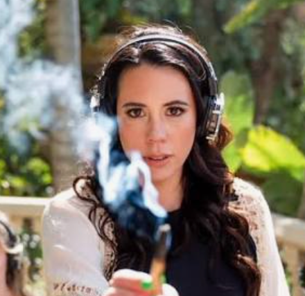 A woman wearing over-ear headphones and a white lace-trim top holds a smoking object in front of her face, with a blurred outdoor background of green foliage.