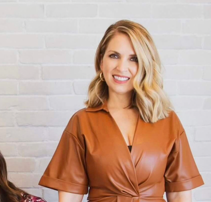 Veronica wearing a brown leather dress against a light brick wall.